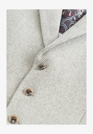 Tessuto beige chiaro con texture e un motivo sottile, caratterizzato da un colletto a scollatura e tre bottoni marroni, con una fodera interna floreale.