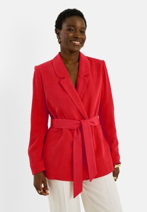Femme souriante, portant un blazer ceinturé rouge vif sur un pantalon blanc, accessoirisée de boucles d'oreilles créoles dorées et de bagues, sur un fond uni.