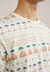 Krótki rękaw kremowa bawełniana koszulka z tropikalnym wzorem przedstawiającym palmy oraz geometryczne motywy w odcieniach zieleni, pomarańczu i niebieskiego.
