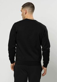 Man met kort haar, van achteren gezien, gekleed in een effen zwarte trui met lange mouwen en een zwarte broek, tegen een grijze achtergrond.