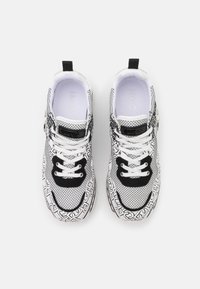 Sneaker in bianco e nero con parte superiore in rete, accenti neri a contrasto e intricato motivo geometrico bianco. Presenta lacci bianchi e un fermacorda nero.