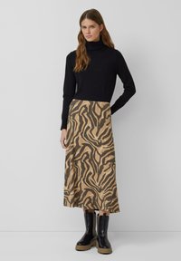 Schwarzer Rollkragenpullover, lockere Passform, kombiniert mit einem hellbraunen Rock mit schwarzem Tiermuster. Schwarze Stiefeletten mit klobigen Sohlen.