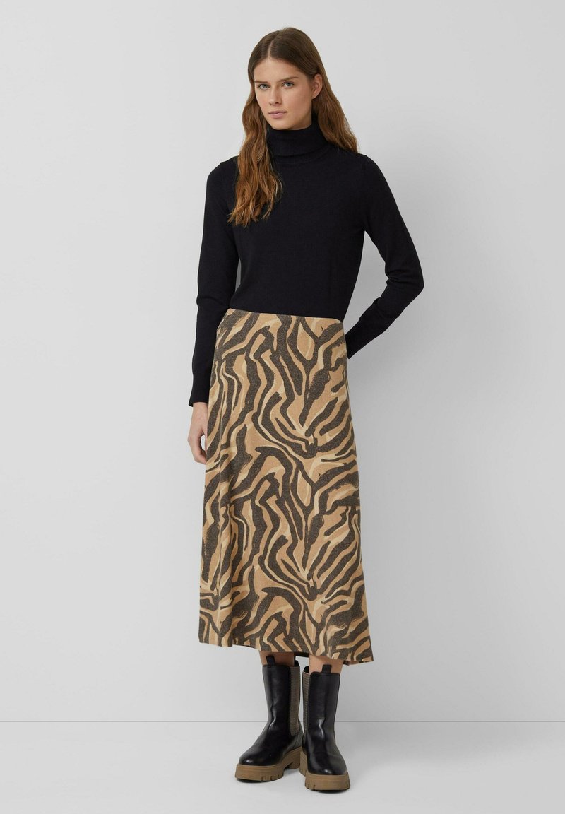 Schwarzer Rollkragenpullover, lockere Passform, kombiniert mit einem hellbraunen Rock mit schwarzem Tiermuster. Schwarze Stiefeletten mit klobigen Sohlen.