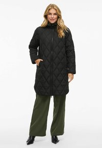 Black quilted long coat with a high collar, front zipper, and side pockets. Features a diamond pattern and a relaxed fit. Worn over green pants.