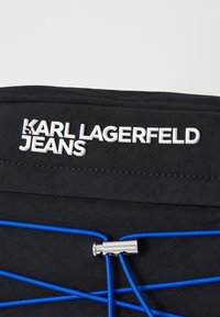 Schwarze Stofftasche mit einem auffälligen weißen Logo "KARL LAGERFELD JEANS" und blauen Gummibanddetails mit einem Metallclip-Akzent.
