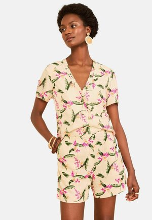 Kvinde iført beige kortærmet romper med blomster, grønne blade og pink blomster, accessoriseret med store runde øreringe og armbånd.