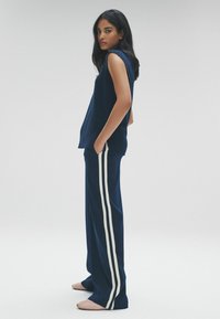 Námornícky modrý pletený top bez rukávov spárovaný s námorníckymi nohavicami s bielymi bočnými pruhmi. Modelka stojí s rukami v vreckách a nosí ploché topánky.