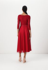 Robe rouge avec un corsage en dentelle à manches trois-quarts, une taille cintrée et une jupe ample et fluide. La jupe présente des plis et une texture lisse.