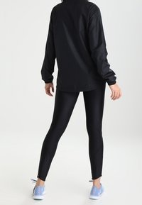 Chaqueta negra y ligera combinada con leggings negros de cintura alta, con una textura suave y un diseño elegante. Zapatillas deportivas azul claro completan el conjunto.