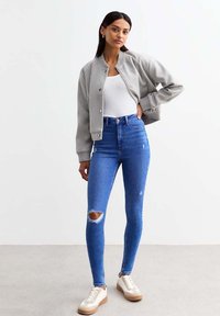 Chaqueta bomber gris con puños acanalados, blusa blanca ajustada, jeans ajustados azul desgastados y zapatillas beige con acentos blancos.