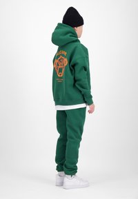 Person wearing green hoodie and pants with orange graphic on the back, white sneakers, and a black beanie, standing sideways on white background.