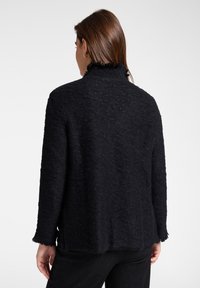 Kobieta o brązowych włosach nosi czarny teksturowany top z długim rękawem z postrzępionymi krawędziami, pokazana od tyłu na jednolitym białym tle.