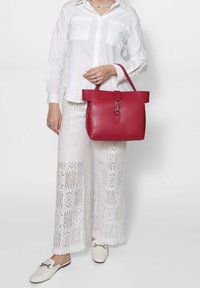 Borsa tote in pelle rossa con una forma strutturata, manici piatti e un dettaglio di chiusura in argento. Indossata con pantaloni di pizzo bianchi e una camicia bianca.