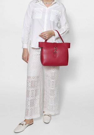 Borsa tote in pelle rossa con una forma strutturata, manici piatti e un dettaglio di chiusura in argento. Indossata con pantaloni di pizzo bianchi e una camicia bianca.