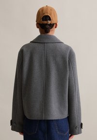 Grijze wolmengeling jasje met brede kraag, verlaagde schouders en knoopmanchetten. Draag het over donkere denim, met een tan kleurige pet op het hoofd.