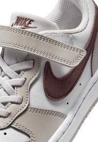 Λευκό και μπεζ αθλητικό παπούτσι Nike με μαρουαν γούνινο σήμα, λευκά κορδόνια και ιμάντα Velcro στο πάνω μέρος.