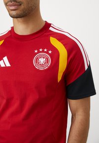 Homem a usar uma camisola de futebol da Alemanha vermelha com riscas brancas, mangas pretas, detalhes amarelos e o logo da Deutscher Fussball-Bund com quatro estrelas.