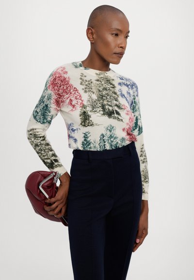 Knit sweater with a cream background featuring a multicolored tree pattern. Worn with dark navy trousers, holding a burgundy clutch.
