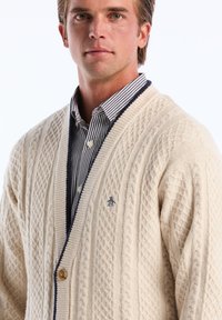 Gebreide crèmekleurige cardigan met een textuurpatroon, voorzien van een marineblauwe kraag en knoop, gedragen over een blauw-witte gestreepte shirt.