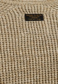 Beige gebreide trui met een textuurpatroon. Zwart label gestikt aan de nek met "FREEDOM UNLIMITED" en "GETEST OP HOGE HOOGTE."