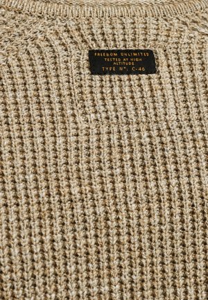 Beige gestrickter Pullover mit einem strukturierten Muster. Schwarzes Etikett auf dem Nacken mit "FREEDOM UNLIMITED" und "GETESTET IN HOHER HÖHE."