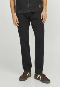 Black denim pants with a straight fit. Worn with brown sneakers featuring white stripes. Material appears sturdy with a smooth texture.