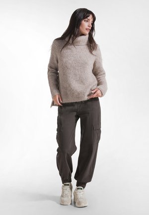 Vrouw draagt een beige coltrui, donkere cargobroek en witte sneakers, staat met handen bij de zakken en kijkt schuin weg.