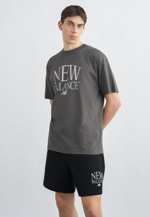 Ung mann iført mørkegrå New Balance t-skjorte og svarte New Balance-shorts, stående med hendene bak ryggen mot en ensfarget bakgrunn.