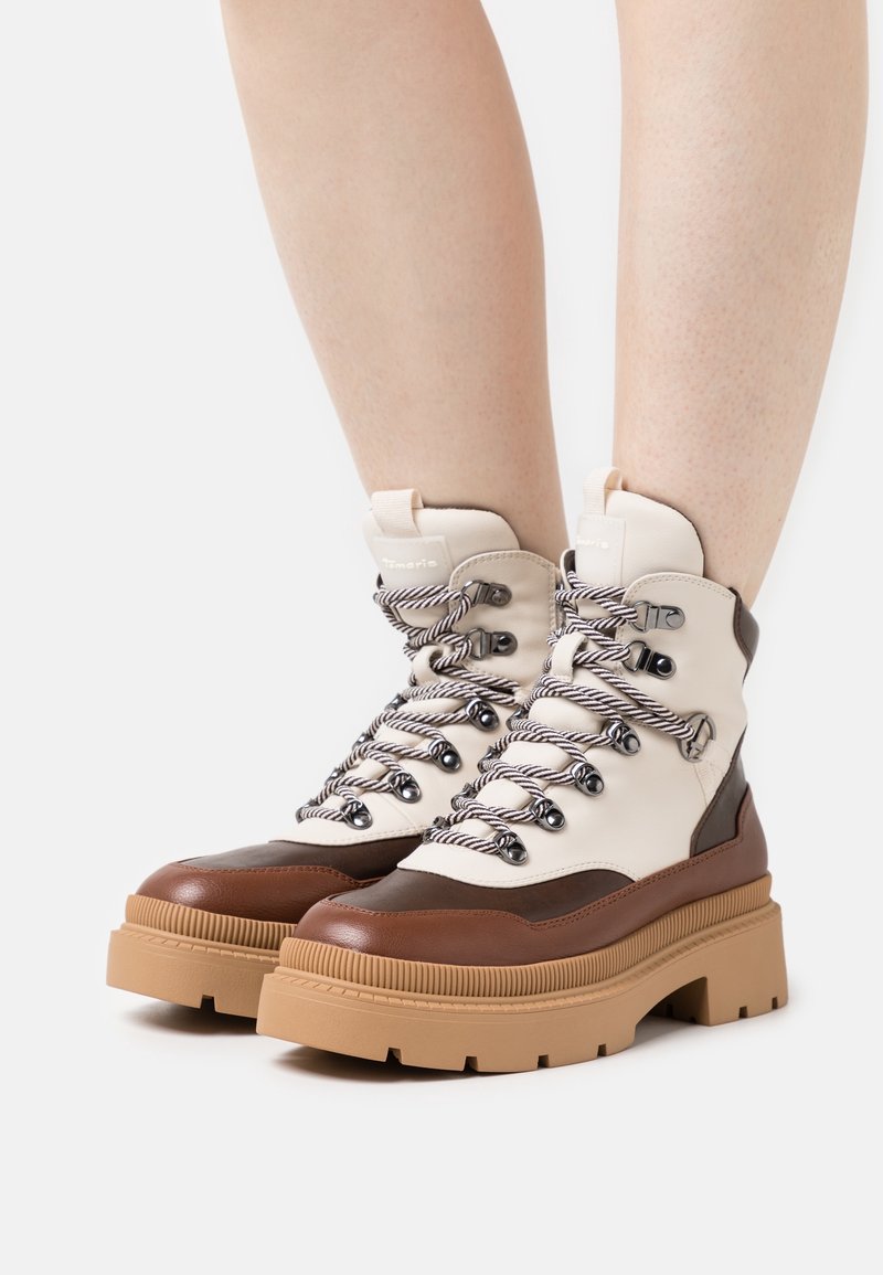Bottes en cuir marron et beige montant jusqu'à la cheville, avec des semelles en caoutchouc épaisses, des lacets noirs et blancs, et des œillets métalliques, portées par une personne aux jambes nues.