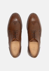 Chaussures habillées en cuir marron avec des détails en brogue, présentant une texture lisse, un système de laçage et une forme de bout légèrement arrondie.