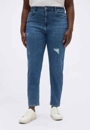 Personne portant un jean taille haute bleu avec de petites zones usées sur la cuisse et le genou, associé à des baskets blanches et un haut blanc.