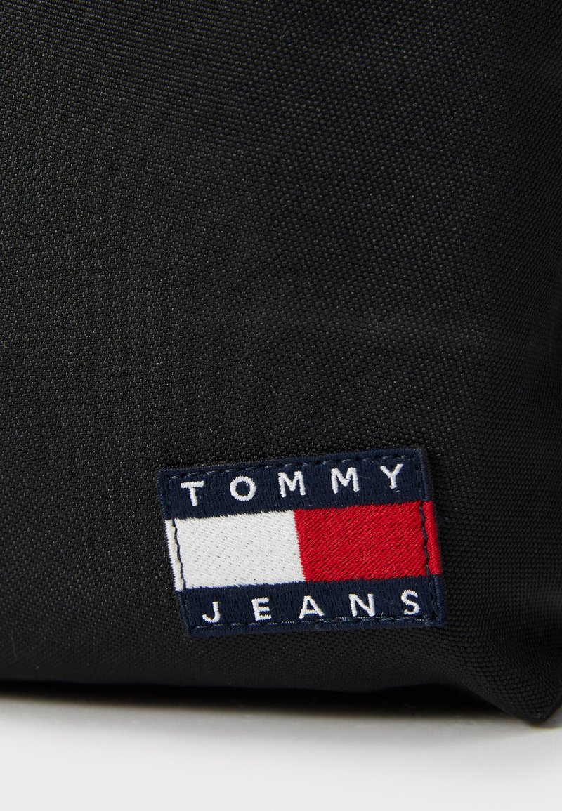 Schwarze Textiltasche mit einem rechteckigen, gestickten Patch mit "TOMMY JEANS" und einem rot-weißen-blauen Logo-Design.