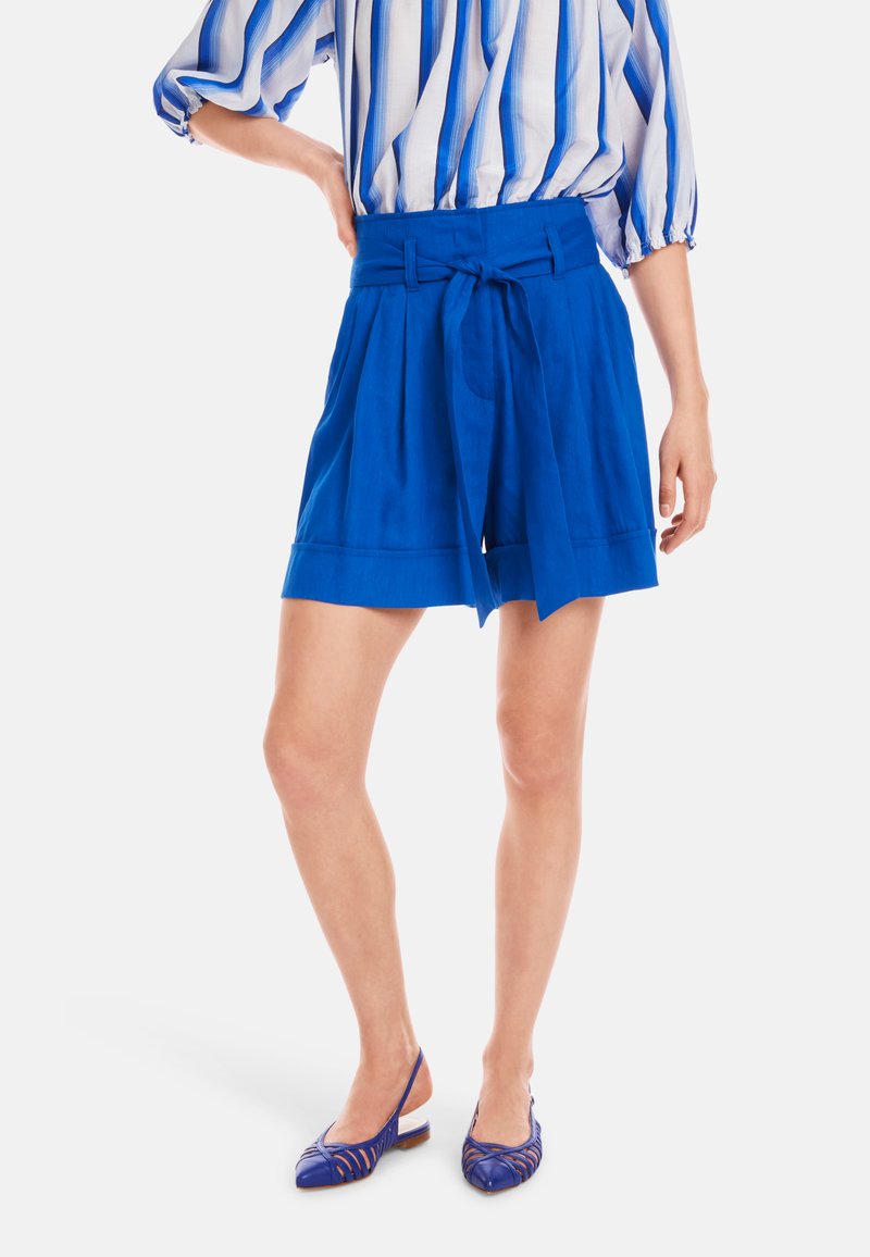 Helle blaue Shorts mit einem taillierten Gürtel und Falten, kombiniert mit blauen Slingback-Schuhen. Der Stoff hat eine glatte Textur und ein strukturiertes Design.