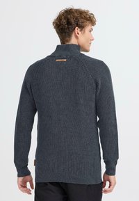 Ciemnoszary sweter z dzianiny z wysokim kołnierzem, ściągaczami i fakturowanym wzorem. Ma małą skórzaną metkę z tyłu.
