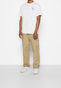 Mies, joka on pukeutunut valkoiseen graafiseen T-paitaan, beigeihin cargo-housuihin ja valkoisiin tennareihin, seisoo yksinkertaista valkoista taustaa vasten.