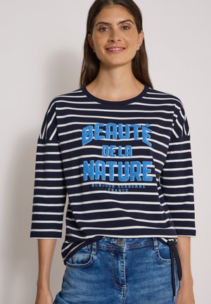 Jeune femme aux longs cheveux bruns porte une chemise rayée bleu marine et blanche avec un texte bleu "Beauté de la Nature" et un jean bleu délavé.