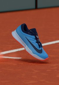 Nike tennissko i blått med mörka accenter, texturerat meshovandel, dämpad sula och livfulla röda detaljer, uppvisad på en lerbana.
