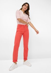 Korallefarbene Jeans mit schlankem Schnitt und umgeschlagenen Säumen, kombiniert mit einem polohemd mit weißen und roten Streifen; Modell trägt weiße Sneakers.