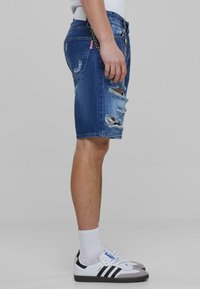 Blauwe denim shorts met een verwasde uitstraling en rafelige uiteinden, vastgemaakt met een riem. Gecombineerd met witte sokken en zwart-witte sneakers.