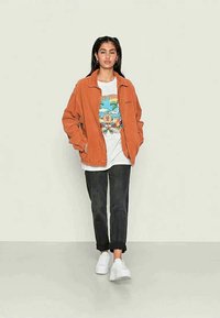 Jaqueta de corduroy laranja, t-shirt branca com design colorido, jeans escuros e ténis brancos. De pé sobre um fundo claro.