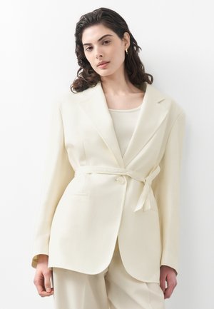 Vrouw met golvend donker haar, gekleed in een off-white blazer met ceintuur over een bijpassende broek en een licht shirt, staand tegen een witte achtergrond.