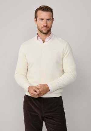 V-Ausschnitt-Pullover aus cremefarbenem Strickstoff, mit gerippten Bündchen und Saum. Getragen über einem rosa Hemd mit Kragen, kombiniert mit braunen Cordhosen.