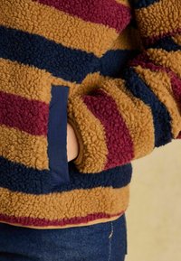 Fleece džemperis svītrotiem toņiem: sinepju dzeltena, bordo un jūras zilā. Ar mīkstu tekstūru un sānu kabatu ar kontrastainu akcentu.