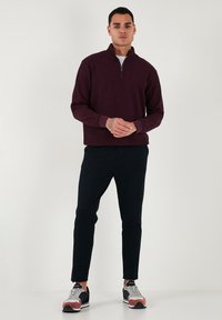 Suéter burdeos con cierre, fabricado en tejido texturizado, pantalones negros ajustados y zapatillas deportivas multicolores con detalles en gris, rojo y azul.