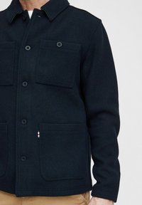 Black textured jacket made of a wool-like fabric; features a collared neck, two chest pockets, and a short buttoned front.