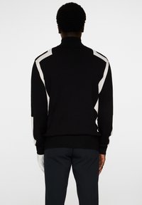 Svart stickad tröja med hög krage och vita accentmönster på axlarna. Tyget ser slätt och strukturerat ut.