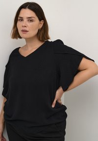 Czarna bluzka o teksturowej powierzchni, w kształcie litery V, z szerokimi rękawami, w połączeniu z ciemnymi spodniami. Prosty fason bez wzorów czy akcentów.