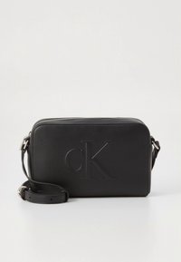 Melna ādas plecu soma ar reljefu "cK" logotipu, taisnstūrveida forma, rāvējslēdzēja aizdare un regulējams siksniņa. Gluda tekstūra ar minimālu dizainu.