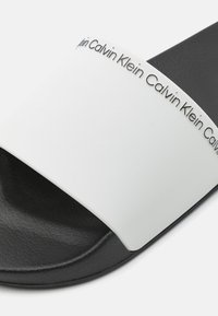 Fekete-fehér csúszó szandál, texturált fekete talppal és sima fehér pánttal, amelyen ismétlődően szerepel a "Calvin Klein" márkaneve.
