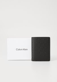 Calvin Klein novčanik u crnoj teksturiranoj koži s suptilnim uzorkom logotipa, uparen s bijelom kutijom s brendom.